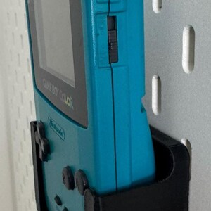 May include: A teal Game Boy Color handheld video game console is mounted on a black wall mount. The console is in a vertical position with the screen facing the right side of the image. The console has a black and white screen and the text "GAME BOY COLOR" is visible on the front of the console.