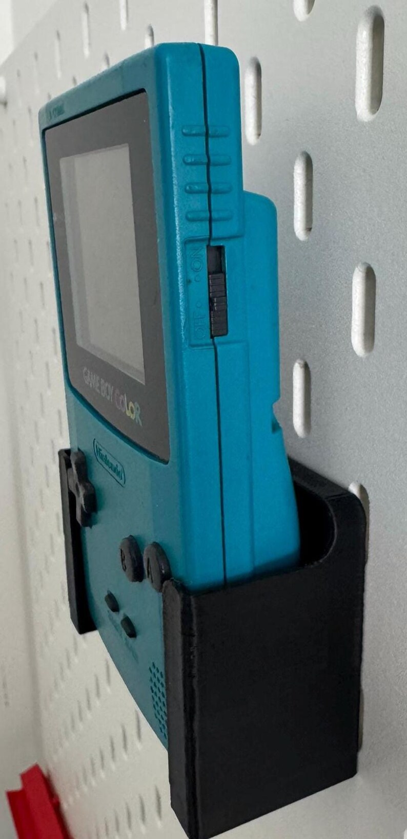 May include: A teal Game Boy Color handheld video game console is mounted on a black wall mount. The console is in a vertical position with the screen facing the right side of the image. The console has a black and white screen and the text "GAME BOY COLOR" is visible on the front of the console.
