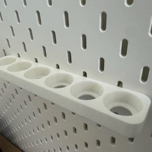May include: A white, 3D-printed pegboard shelf with six circular openings. The shelf is mounted on a white pegboard with evenly spaced holes. The design is simple and functional, suitable for organizing small items.