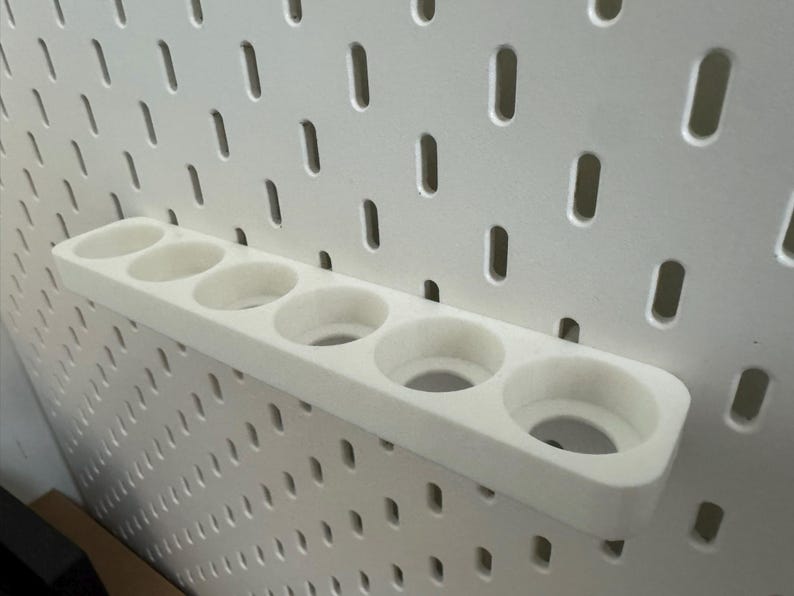 May include: A white, 3D-printed pegboard shelf with six circular openings. The shelf is mounted on a white pegboard with evenly spaced holes. The design is simple and functional, suitable for organizing small items.