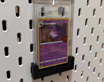 May include: A Gengar Pokémon trading card, graded and encased in a clear plastic holder, mounted on a black display stand. The card features a purple Gengar illustration and the text "Gengar". The holder is attached to a white pegboard.