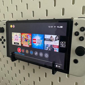 May include: A white Nintendo Switch console with a black screen displaying the game menu. The console is mounted on a white pegboard wall. The screen shows several game icons, including "Big Brain", "Super Mario Odyssey", "LEGO City Undercover", "Assassin's Creed: The Rebel Collection", and "Assassin's Creed: Rogue". The time is 12:39 and the battery is at 94%.