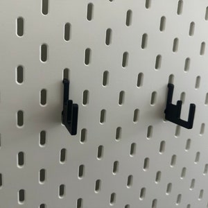 May include: Two black plastic pegboard hooks mounted on a white pegboard wall.