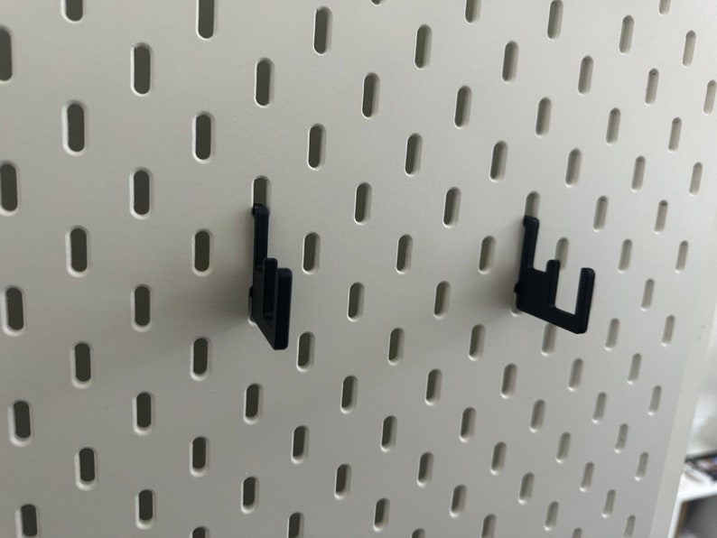 May include: Two black plastic pegboard hooks mounted on a white pegboard wall.