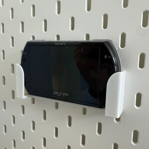 Pu&ograve; includere: Una console di gioco portatile Sony PSP nera montata su una parete forata bianca con un supporto bianco. La PSP &egrave; centrata, mostrando il logo Sony e il marchio PSP. La parete forata ha fessure verticali distanziate uniformemente.