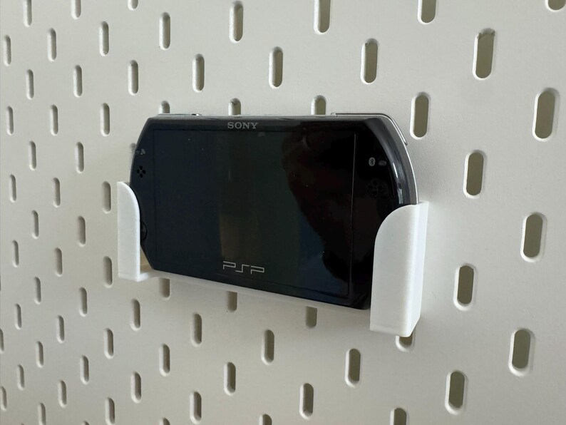 Pu&ograve; includere: Una console di gioco portatile Sony PSP nera montata su una parete forata bianca con un supporto bianco. La PSP &egrave; centrata, mostrando il logo Sony e il marchio PSP. La parete forata ha fessure verticali distanziate uniformemente.