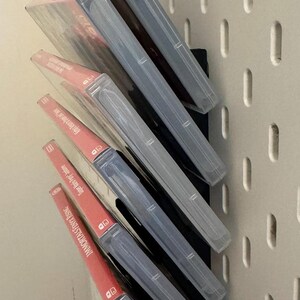 Pu&ograve; includere: Un rack di stoccaggio per videogiochi a parete con sei custodie per videogiochi. Le custodie sono in plastica trasparente con etichette rosse e bianche. Il rack &egrave; nero e ha uno sfondo bianco.