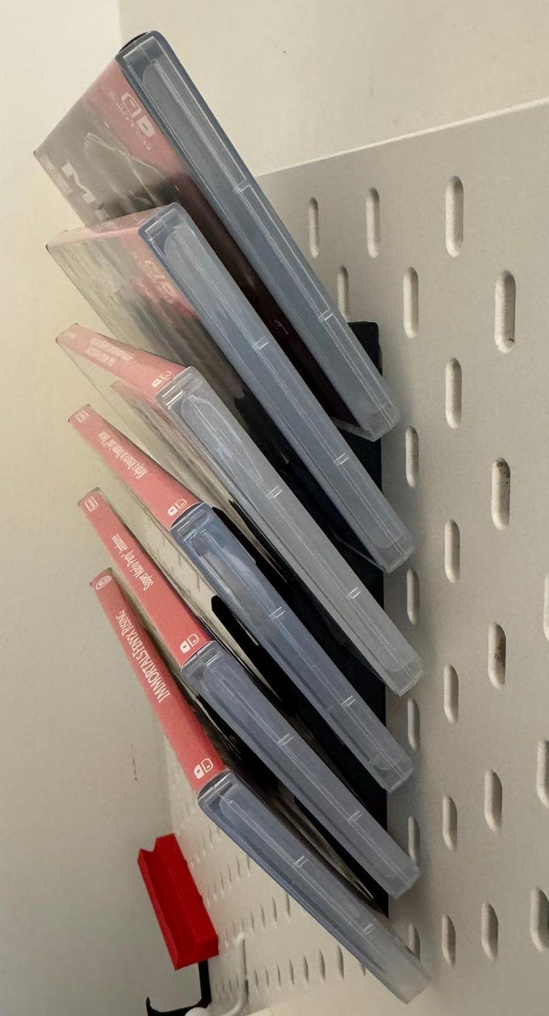 Pu&ograve; includere: Un rack di stoccaggio per videogiochi a parete con sei custodie per videogiochi. Le custodie sono in plastica trasparente con etichette rosse e bianche. Il rack &egrave; nero e ha uno sfondo bianco.