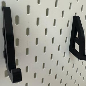 May include: Two black plastic wall mounted hooks, designed to hold items like hats, bags, or other accessories. The hooks are mounted on a white pegboard.