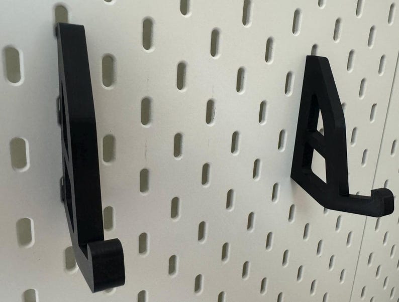 May include: Two black plastic wall mounted hooks, designed to hold items like hats, bags, or other accessories. The hooks are mounted on a white pegboard.
