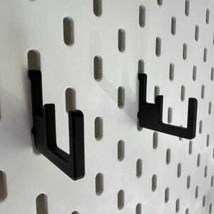 Puede incluir: Dos ganchos de metal negro montados en una pared perforada blanca. Los ganchos en forma de L est&aacute;n dise&ntilde;ados para colgar objetos. La pared perforada tiene agujeros ovalados espaciados uniformemente. Los ganchos tienen un dise&ntilde;o simple y moderno.