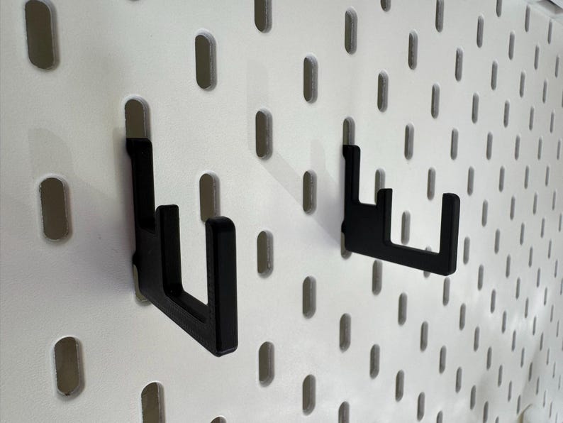 Puede incluir: Dos ganchos de metal negro montados en una pared perforada blanca. Los ganchos en forma de L est&aacute;n dise&ntilde;ados para colgar objetos. La pared perforada tiene agujeros ovalados espaciados uniformemente. Los ganchos tienen un dise&ntilde;o simple y moderno.