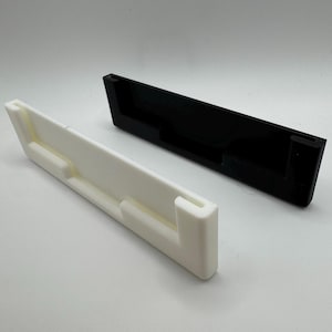 以下が含まれることがあります： 白と黒の2つの3Dプリントされた壁掛け。どちらも長方形で、中央に電話やその他のデバイスを保持するためのスロットがあります。