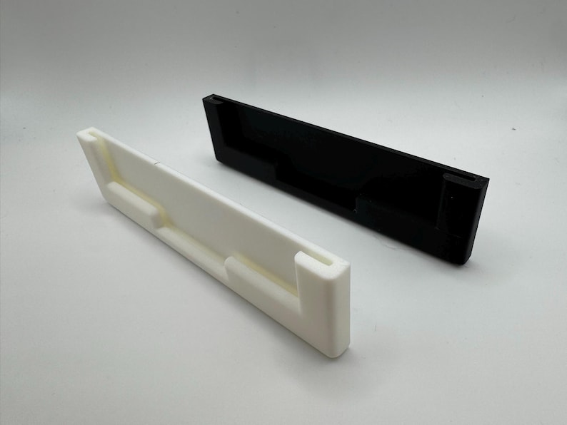 以下が含まれることがあります： 白と黒の2つの3Dプリントされた壁掛け。どちらも長方形で、中央に電話やその他のデバイスを保持するためのスロットがあります。