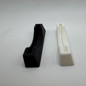 Puede incluir: Dos soportes impresos en 3D, uno negro y otro blanco, se muestran sobre un fondo blanco. El soporte negro tiene un dise&ntilde;o curvo, mientras que el blanco es rectangular con una ranura.