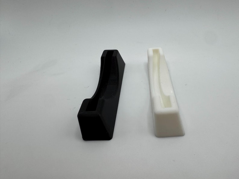Puede incluir: Dos soportes impresos en 3D, uno negro y otro blanco, se muestran sobre un fondo blanco. El soporte negro tiene un dise&ntilde;o curvo, mientras que el blanco es rectangular con una ranura.