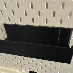 Peut inclure: &Eacute;tag&egrave;re de rangement noire, rectangulaire, avec des c&ocirc;t&eacute;s inclin&eacute;s, fix&eacute;e sur un panneau perfor&eacute; blanc. L'&eacute;tag&egrave;re est con&ccedil;ue pour contenir des objets et est faite d'un mat&eacute;riau sombre.