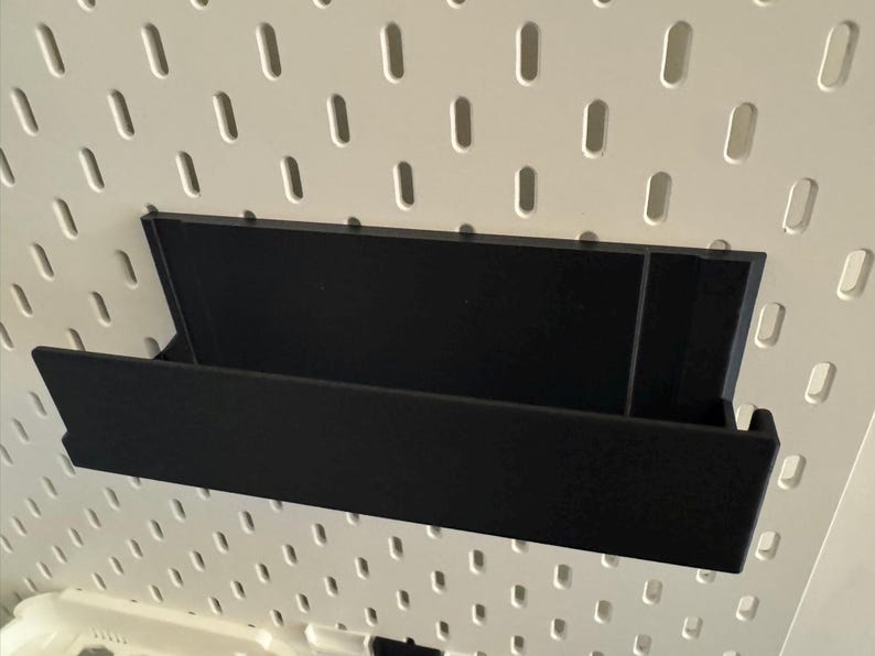 Peut inclure: &Eacute;tag&egrave;re de rangement noire, rectangulaire, avec des c&ocirc;t&eacute;s inclin&eacute;s, fix&eacute;e sur un panneau perfor&eacute; blanc. L'&eacute;tag&egrave;re est con&ccedil;ue pour contenir des objets et est faite d'un mat&eacute;riau sombre.