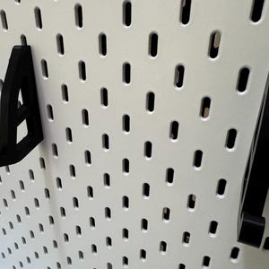 May include: Black pegboard hooks in various shapes are displayed against a white pegboard background. The hooks are designed to fit into the pegboard holes, providing a storage or display solution. The image showcases the hooks' design and functionality.