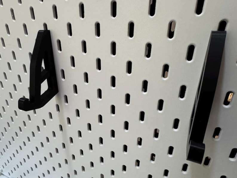 May include: Black pegboard hooks in various shapes are displayed against a white pegboard background. The hooks are designed to fit into the pegboard holes, providing a storage or display solution. The image showcases the hooks' design and functionality.