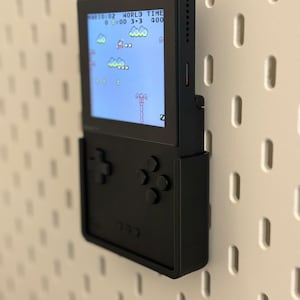 Op de afbeelding: Een zwarte draagbare videogameconsole met een scherm waarop een gepixelde videogame wordt weergegeven. De console heeft een directionele pad en knoppen. De console is gemonteerd op een witte pegboardwand.