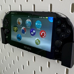 Op de afbeelding: Een zwarte Sony PlayStation Vita handheld-gameconsole met een zwarte wandbevestiging. Het scherm van de console toont het startmenu met pictogrammen voor Welcome Park, PS Store, Browser, Berichten, Party en Ouderlijk toezicht. Het scherm toont ook de tijd 0:00 en 95% batterijduur.