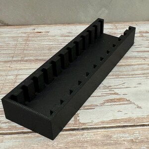 Puede incluir: Un organizador de herramientas rectangular negro con m&uacute;ltiples ranuras verticales y recortes triangulares. El organizador est&aacute; hecho de un material s&oacute;lido y se encuentra sobre una superficie de madera. El dise&ntilde;o sugiere que es para guardar herramientas o objetos peque&ntilde;os.