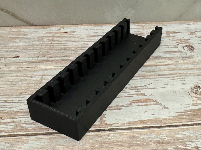 Puede incluir: Un organizador de herramientas rectangular negro con m&uacute;ltiples ranuras verticales y recortes triangulares. El organizador est&aacute; hecho de un material s&oacute;lido y se encuentra sobre una superficie de madera. El dise&ntilde;o sugiere que es para guardar herramientas o objetos peque&ntilde;os.