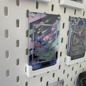 K&ouml;nnte beinhalten: Eine wei&szlig;e Lochwand mit mehreren durchsichtigen Plastik-Kartenhaltern. Die Kartenhalter halten verschiedene Pok&eacute;mon-Sammelkarten, darunter eine Karte mit einem rosa und wei&szlig;en Pok&eacute;mon namens "Minccino".