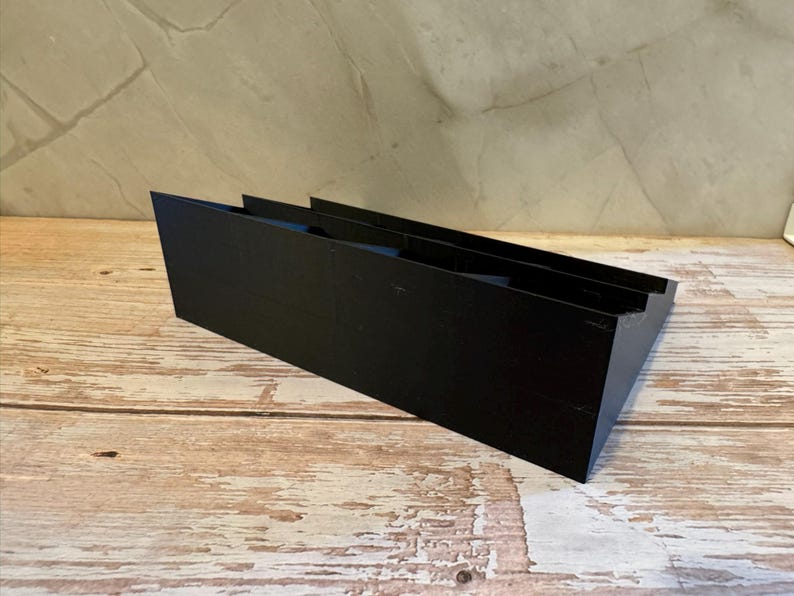 Peut inclure: Un objet rectangulaire noir et inclin&eacute; avec deux rainures parall&egrave;les. L'objet est fait d'un mat&eacute;riau solide et repose sur une surface en bois. L'arri&egrave;re-plan est un mur de couleur claire.