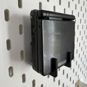 May include: A black Nintendo Game Boy Advance SP handheld console mounted on a white pegboard. The console is in a black holder with the Nintendo logo visible. The pegboard has evenly spaced oval holes.