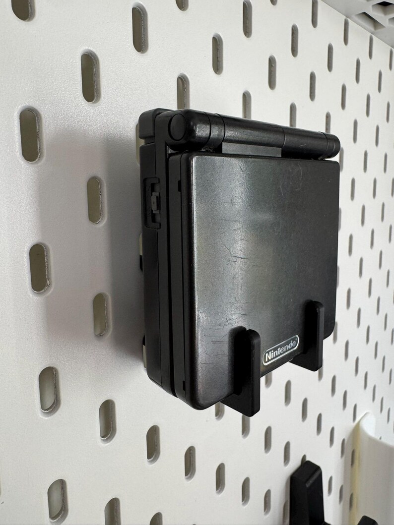 May include: A black Nintendo Game Boy Advance SP handheld console mounted on a white pegboard. The console is in a black holder with the Nintendo logo visible. The pegboard has evenly spaced oval holes.