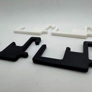 May include: Four plastic brackets in a studio shot. Two are black, and two are white. Each bracket has a rectangular base with a hook extending from one side. The brackets are designed to hold or support something.