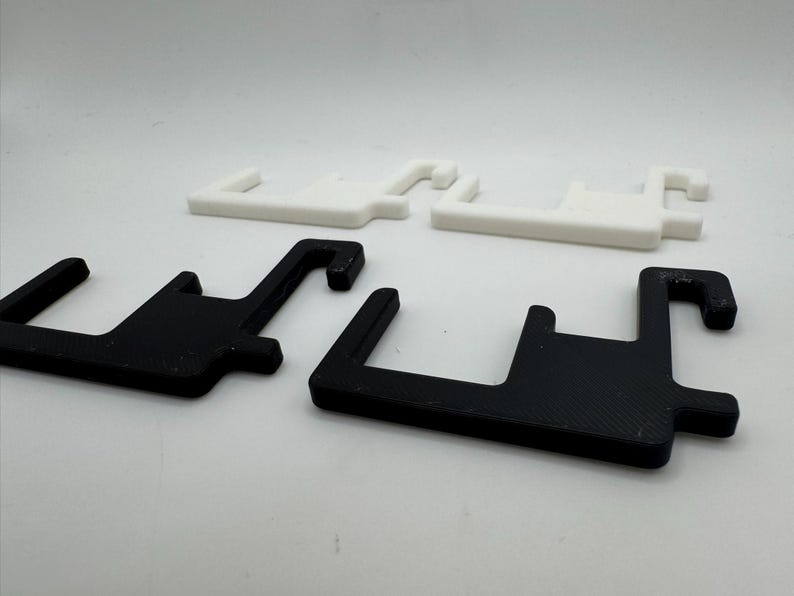 May include: Four plastic brackets in a studio shot. Two are black, and two are white. Each bracket has a rectangular base with a hook extending from one side. The brackets are designed to hold or support something.