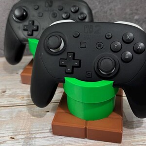 K&ouml;nnte beinhalten: Zwei schwarze Gamecontroller mit wei&szlig;en Akzenten, die auf gr&uuml;nen und braunen St&auml;ndern ruhen. Die Controller haben mehrere Tasten und Richtungspads. Die St&auml;nder sind als gr&uuml;ne R&ouml;hre auf einem braunen Block gestaltet.