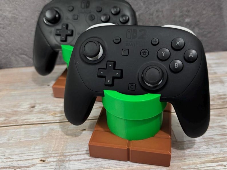 K&ouml;nnte beinhalten: Zwei schwarze Gamecontroller mit wei&szlig;en Akzenten, die auf gr&uuml;nen und braunen St&auml;ndern ruhen. Die Controller haben mehrere Tasten und Richtungspads. Die St&auml;nder sind als gr&uuml;ne R&ouml;hre auf einem braunen Block gestaltet.