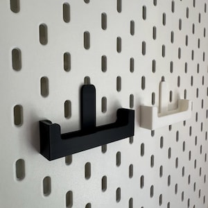 May include: Black and white pegboard hooks. The black hook is mounted on a white pegboard. A white hook is also mounted on the pegboard. The hooks are designed to hold items. The pegboard has evenly spaced oval holes.