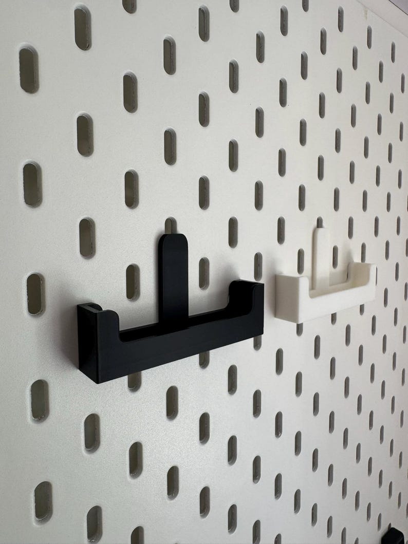 May include: Black and white pegboard hooks. The black hook is mounted on a white pegboard. A white hook is also mounted on the pegboard. The hooks are designed to hold items. The pegboard has evenly spaced oval holes.