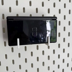 Puede incluir: Una consola de juegos port&aacute;til Nintendo DS Lite negra est&aacute; montada en una pared perforada blanca con soportes de pl&aacute;stico blanco. La consola es rectangular, con un acabado brillante y un peque&ntilde;o bot&oacute;n de encendido.