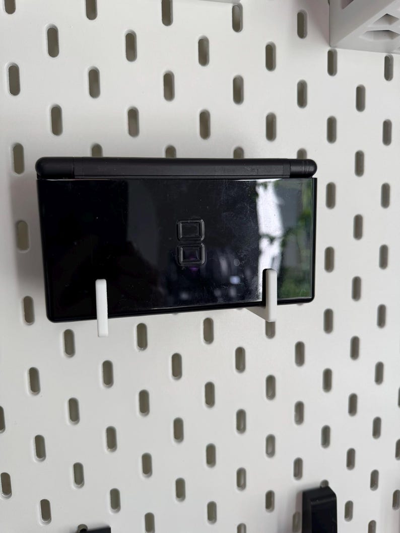 Puede incluir: Una consola de juegos port&aacute;til Nintendo DS Lite negra est&aacute; montada en una pared perforada blanca con soportes de pl&aacute;stico blanco. La consola es rectangular, con un acabado brillante y un peque&ntilde;o bot&oacute;n de encendido.