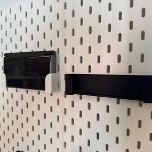 May include: A white pegboard with a black rectangular shelf and a black electronic device in a white holder. The pegboard has evenly spaced oval holes.