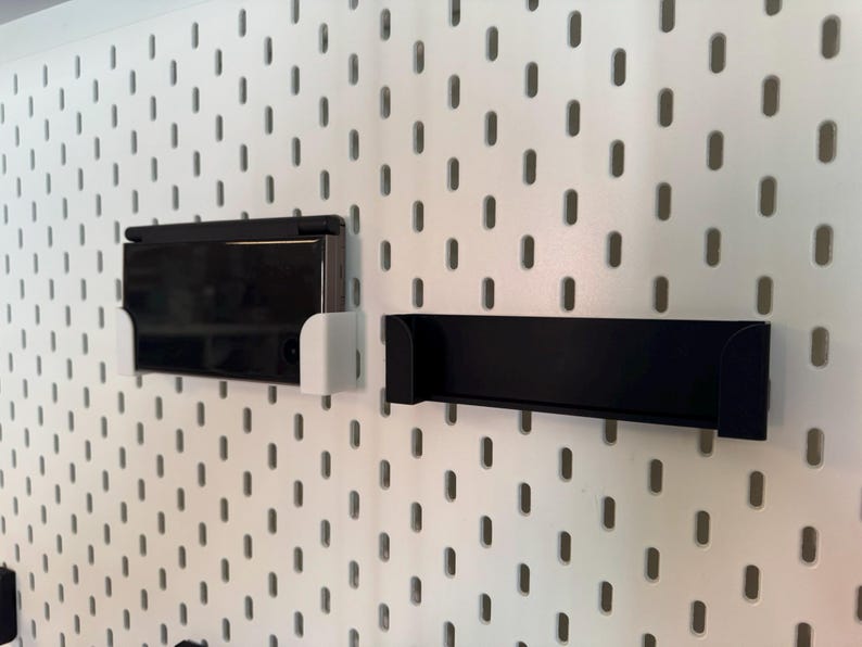 May include: A white pegboard with a black rectangular shelf and a black electronic device in a white holder. The pegboard has evenly spaced oval holes.
