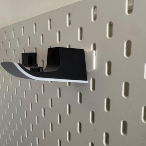 K&ouml;nnte beinhalten: Ein schwarz-wei&szlig;es Wandregal, das zur Aufnahme eines Gegenstandes bestimmt ist, ist an einer wei&szlig;en Lochwand befestigt. Das Regal hat ein geschwungenes Design mit wei&szlig;em Rand. Die Lochwand hat eine Reihe von gleichm&auml;&szlig;ig verteilten, vertikalen ovalen L&ouml;chern.