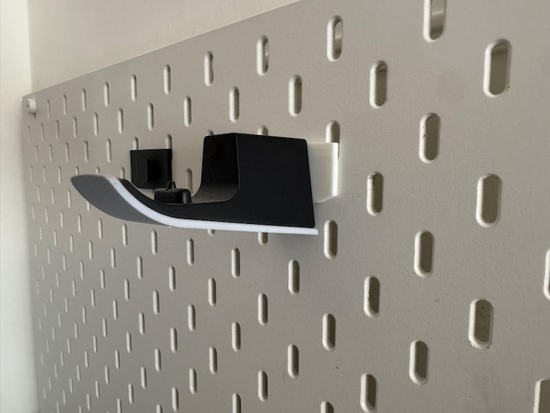 K&ouml;nnte beinhalten: Ein schwarz-wei&szlig;es Wandregal, das zur Aufnahme eines Gegenstandes bestimmt ist, ist an einer wei&szlig;en Lochwand befestigt. Das Regal hat ein geschwungenes Design mit wei&szlig;em Rand. Die Lochwand hat eine Reihe von gleichm&auml;&szlig;ig verteilten, vertikalen ovalen L&ouml;chern.