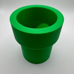 May include: A bright green, cylindrical object resembling a pipe, with a wide opening at the top and a slightly narrower base. The pipe is a solid, vibrant green color and appears to be made of a smooth material. The design is reminiscent of a video game.