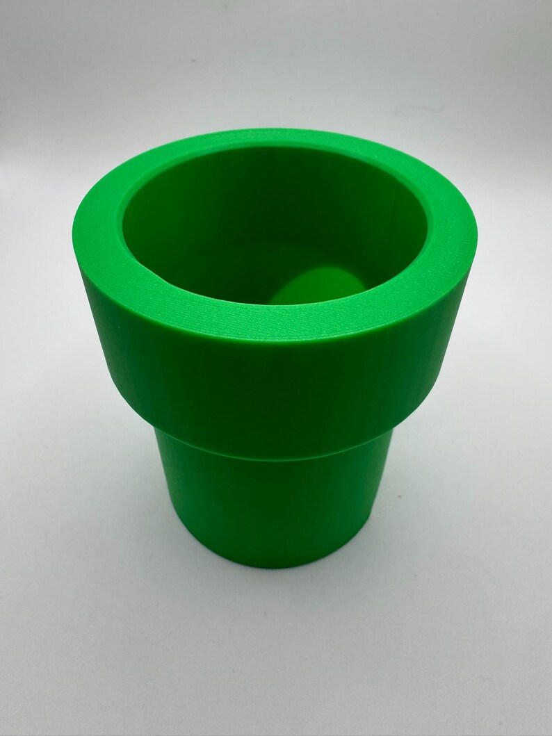 May include: A bright green, cylindrical object resembling a pipe, with a wide opening at the top and a slightly narrower base. The pipe is a solid, vibrant green color and appears to be made of a smooth material. The design is reminiscent of a video game.