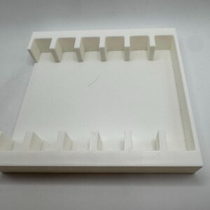May include: A white, rectangular organizer with multiple vertical slots. The organizer has a raised border and is designed to hold small items. The slots are evenly spaced and appear to be of uniform size.