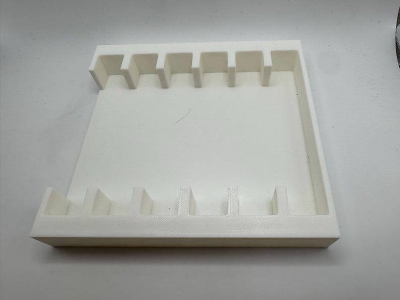 May include: A white, rectangular organizer with multiple vertical slots. The organizer has a raised border and is designed to hold small items. The slots are evenly spaced and appear to be of uniform size.
