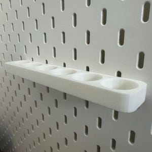 May include: A white, 3D-printed pegboard shelf with five circular openings. The shelf is mounted on a white pegboard with evenly spaced, oblong holes. The shelf is designed to hold small items and is made of a smooth, solid material.