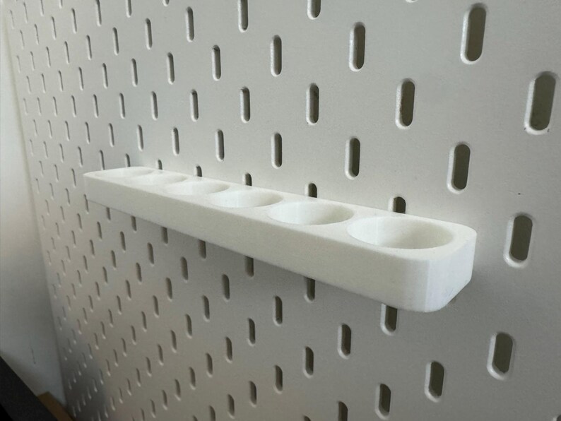 May include: A white, 3D-printed pegboard shelf with five circular openings. The shelf is mounted on a white pegboard with evenly spaced, oblong holes. The shelf is designed to hold small items and is made of a smooth, solid material.